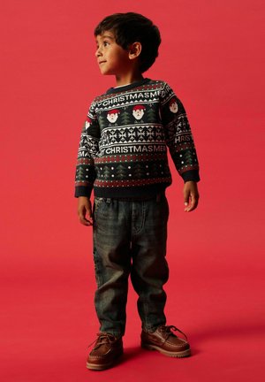 Kind in een zwarte kersttrui met patronen van de kerstman en sneeuwvlokken, gecombineerd met blauw denim jeans en bruine veterschoenen.