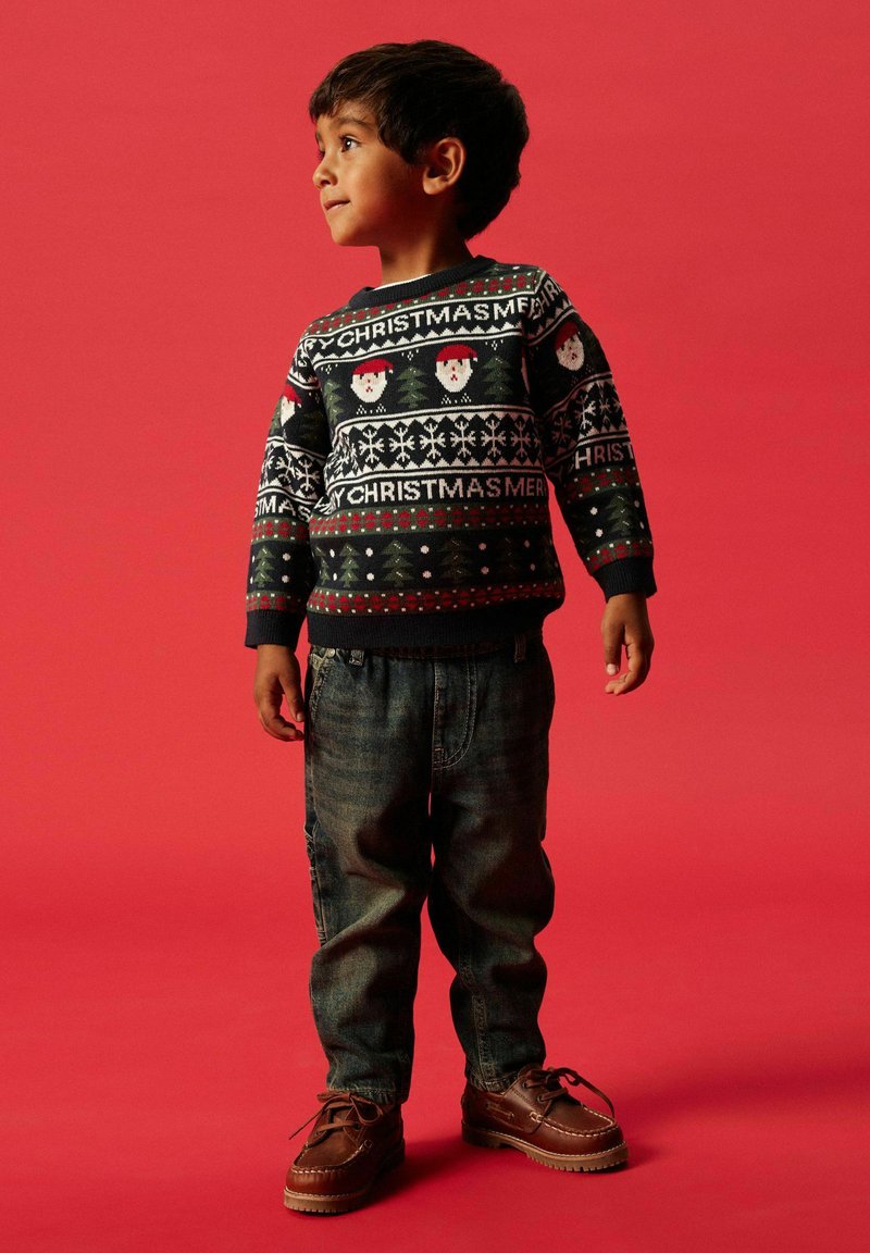 Bambino che indossa un maglione di Natale nero con motivi di Babbo Natale e fiocchi di neve, abbinato a jeans blu in denim e scarpe marroni con lacci.
