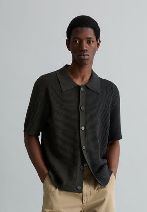 Młody mężczyzna w czarnej, teksturowanej, krótkorękawowej koszuli zapinanej na guziki z kołnierzykiem, stojący z rękami w kieszeniach beżowych spodni na jednolitym tle.