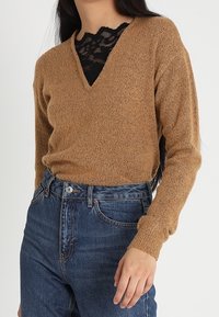 Maglione marrone a maglia con scollo a V e maniche lunghe, sovrapposto a un top in pizzo nero. Caratterizzato da un tessuto strutturato e una vestibilità rilassata. Indossato con jeans blu a vita alta.