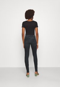 Top negro de canalé combinado con jeans ajustados oscuros. La persona lleva sandalias de tacón verdes. El fondo es blanco liso.