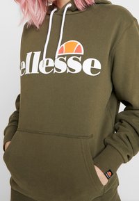 Olivgrön hoodie med vitt "ellesse"-logotyp, dragsko på huvan, kängurufickor och ribbade mudd. Har en liten flerfärgad märke.