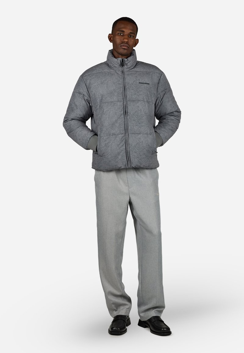 Hombre de pie con las manos en los bolsillos, vistiendo una chaqueta acolchada gris, pantalones gris claro y zapatos de vestir negros sobre un fondo blanco.