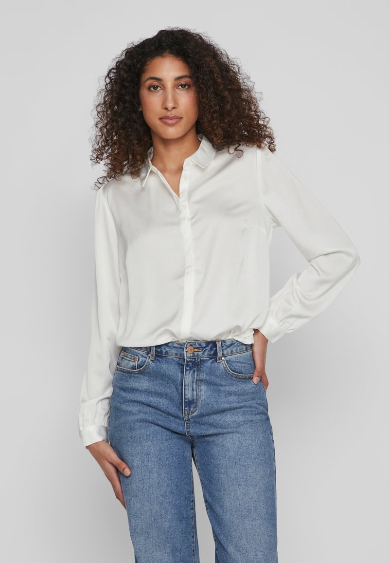 Camicia bianca con bottoni, realizzata in tessuto liscio, a maniche lunghe, abbinata a jeans in denim blu chiaro, con una vestibilità classica e tasche frontali.