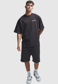 Camiseta de algodón negra de corte oversized con cuello redondo, combinada con pantalones cortos negros a juego. Presenta un sencillo detalle del logo. Lleva puestas zapatillas blancas.
