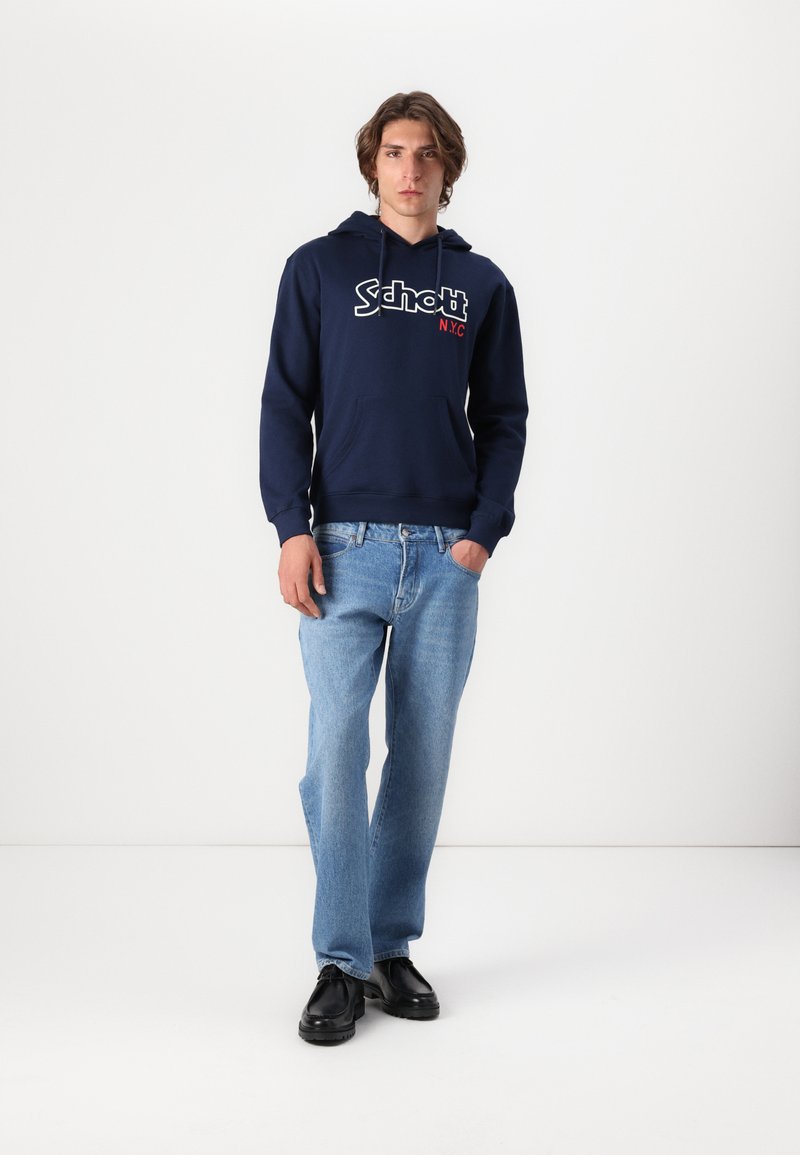 Haut à capuche bleu marine avec un logo blanc et rouge, poche avant, poignets côtelés, associé à un jean bleu clair et des chaussures noires. Design simple et décontracté.