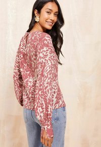 Blusa a maniche lunghe in rosa e crema con stampa animale astratta, dotata di apertura a chiave sul retro e polsini annodati, indossata con jeans blu.