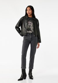 Giacca corta nera in similpelle con colletto, indossata sopra una t-shirt grafica, abbinata a jeans grigi a vita alta e stivaletti neri.