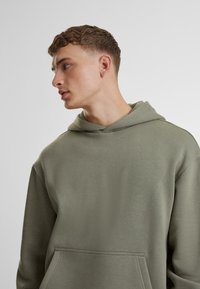 Sudadera verde oliva con un bolsillo frontal, capucha con cordón y un acabado suave y texturizado. Corte relajado con un diseño minimalista.