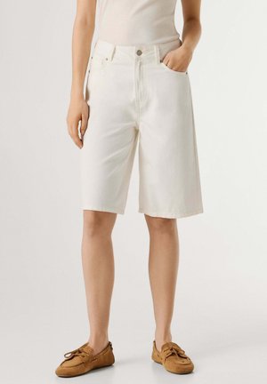 Person trägt weiße, knielange Shorts mit hohem Bund, beige ärmelloses Oberteil und braune Mokassin-Schnürschuhe, steht mit einer Hand in der Tasche.