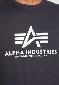 Tmavě šedé tričko vyrobené z bavlny, s velkým bílým grafickým logem s písmenem 'A' a horizontálními pruhy, text zní "ALPHA INDUSTRIES."