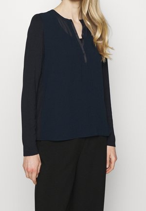 Woman wearing a long-sleeve black top with navy blue sleeveless overlay and black pants, standing against a plain white background.