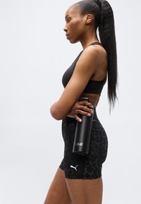 Γυναίκα σε φόρμα γυμναστικής μαύρου χρώματος, κρατώντας μαύρο μπουκάλι νερού Casall, στέκεται σε προφίλ με σταυρωμένα τα χέρια της πάνω σε απλό λευκό φόντο.