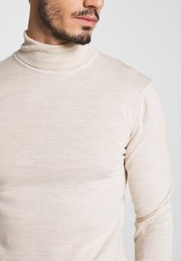 Beige Rollkragenpullover aus leichtem Strickstoff. Mit enganliegenden langen Ärmeln und einem engen, gerippten Halsausschnitt für ein sauberes Finish.