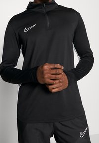 Black, long-sleeve Nike top with a zip mock collar; features white side accents and a white logo on the chest; smooth fabric texture.