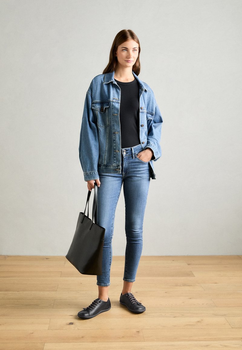 Jeansjacka över en svart skjorta, tillsammans med blå tajta jeans och svarta sneakers. Håller en svart tote-väska. Bakgrund av trägolv.