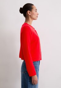 Femme avec les cheveux en chignon, portant un cardigan rouge vif à boutons et une jupe en denim bleue, se tenant de côté contre un fond blanc uni.
