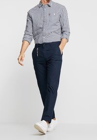 Uomo che cammina indossando una camicia a scacchi bianca e nera, pantaloni blu scuro con un portachiavi e sneaker bianche su uno sfondo grigio uniforme.