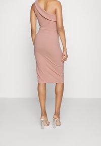 Femme portant une robe midi ajustée sans manches de couleur rose poudré avec un décolleté asymétrique et des sandales à talons clairs, se tenant sur un sol gris.