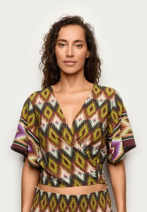 Femme aux cheveux bouclés portant un haut cache-cœur à motif coloré et à col en V, ainsi qu'une jupe assortie, debout sur un fond blanc uni.