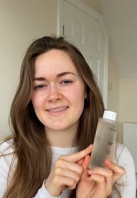 Bouteille transparente avec un bouchon blanc, étiquetée "Pureté du Printemps des Roses". La bouteille a un design minimaliste et est tenue par une personne aux longs cheveux.