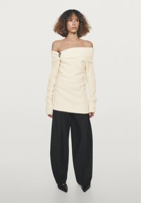 Pull crème tricoté à épaules dénudées avec un détail de broche, associé à un pantalon noir à jambes larges et des chaussures noires pointues. Texture lisse, coupe décontractée.