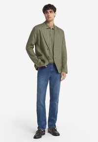 Olivengrüner Leinenblazer über einem passenden Polo, kombiniert mit blauen Jeans und dunklen Lederschuhen mit braunen Schnürsenkeln. Schlichte, taillierte Passform.