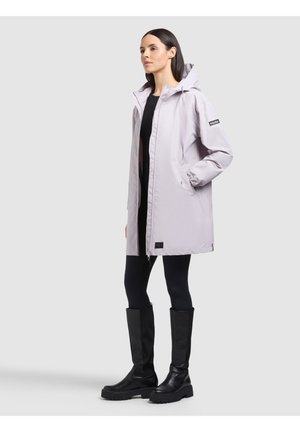 Žena v svetlosivej kapucňovej bunde, čiernych legínach a čiernych čižmách po kolená stojí pred jednoduchým svetlosivým pozadím.