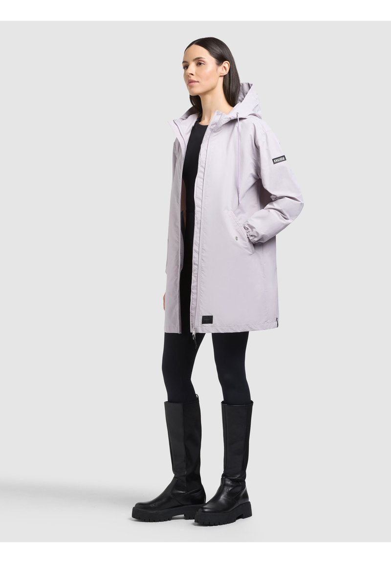 Femme portant une veste à capuche gris clair, des leggings noirs et des bottes noires montantes, debout devant un fond uni gris clair.