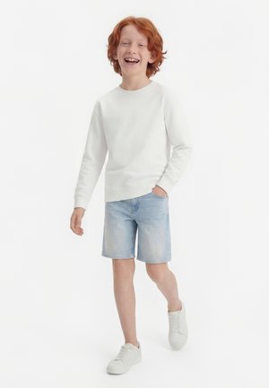 Vörös hajú fiú sétál és mosolyog, fehér hosszú ujjú inget, világos farmer rövidnadrágot és fehér tornacipőt viselve, fehér háttér előtt.