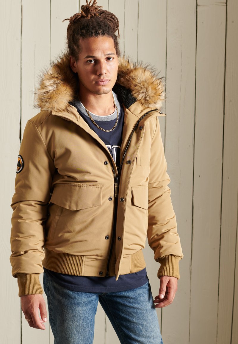 Chaqueta acolchada color beige con capucha forrada de piel, múltiples bolsillos frontales y puños acanalados. Llevada sobre una camiseta azul marino y jeans azules.