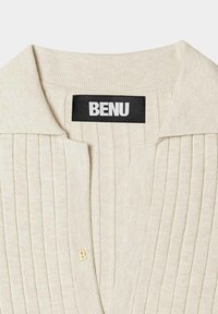 Kremowy sweter z dzianiny z prążkowaną fakturą, miękkim kołnierzem i złotym akcentem w kształcie litery 'B' w pobliżu zapięcia. Wykonany z lekkiego materiału.