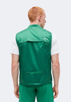 Mężczyzna z krótkimi czerwonymi włosami, ubrany w zieloną kamizelkę bez rękawów na białej koszuli, zwrócony tyłem, pokazując tył kamizelki z małą kieszonką na zamek i logo "BORG".