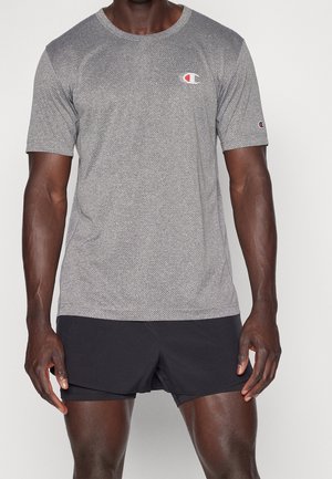 Man draagt een grijs gestructureerd Champion T-shirt met korte mouwen en zwarte sportshorts, staand tegen een effen witte achtergrond.