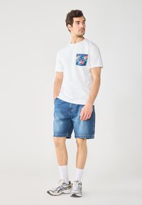 White t-shirt with a blue floral pocket, paired with denim shorts. Model wears gray and black sneakers, white crew socks. Neutral background.