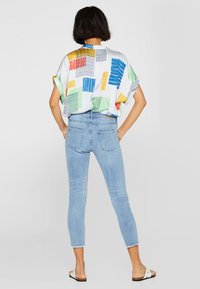 Välj en lös kortärmad skjorta med färgglada geometriska mönster på en ljus bakgrund, kombinerad med ljusblå tajta jeans och vita sandaler.