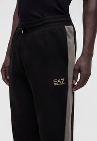 Άνδρας που φοράει μαύρη και γκρι φόρμα EA7 Emporio Armani με κορδόνι στη μέση και χρυσλό λογότυπο στον αριστερό μηρό.