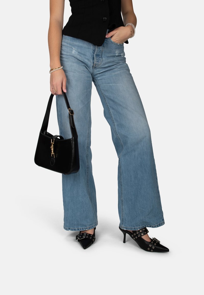 Donna che indossa jeans azzurri a gamba larga, top nero senza maniche, scarpe nere con tacchi alti e fibbie, e tiene una borsa nera nella mano sinistra.