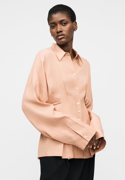 Γυναίκα με κοντά μαλλιά που φοράει μια υπερμεγέθη πουκάμισο ροδακινί με κουμπιά και μαύρο παντελόνι, ποζάρει μπροστά σε ένα απλό φωτεινό φόντο.