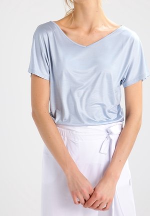 Femme portant un t-shirt à manches courtes bleu clair avec un col en V, rentré dans une jupe portefeuille blanche nouée à la taille, debout les mains jointes.