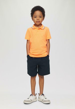 Orange Polohemd mit Kragen, kombiniert mit dunklen Shorts. Das Kind trägt weiße und graue Sneakers mit schwarzen Akzenten. Neutraler Hintergrund.