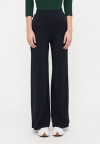 Black wide-leg ribbed pants with an elastic waistband, paired with white sneakers. Fabric shows vertical texture and soft finish.