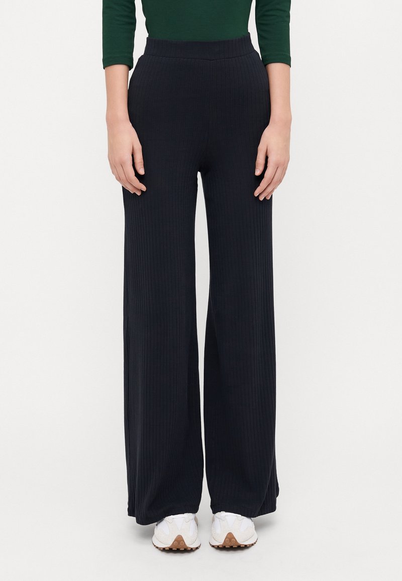 Black wide-leg ribbed pants with an elastic waistband, paired with white sneakers. Fabric shows vertical texture and soft finish.