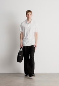 Bílá polokošile s krátkými rukávy a dvěma knoflíky, spárovaná s černými, volnými kalhotami. Modelka drží černou tašku s rukojetí.