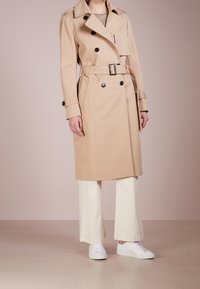 Trench coat beige con design doppiopetto, ampio colletto, vita con cintura e dettagli con bottoni. Abbinato a pantaloni larghi crema e sneakers bianche.