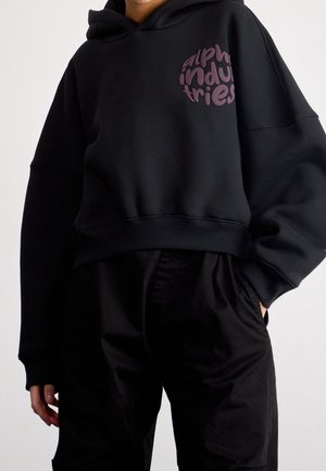 Personne portant un sweat à capuche court noir avec le texte "alpha industries" sur la poitrine et un pantalon noir, main dans la poche, sur un fond uni.