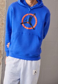 Blauer Hoodie mit einer Fronttasche und Kordelzugkapuze, der eine schwarze Silhouette und eine orangefarbene Grafik in einem kreisförmigen Design zeigt. Weiße Jogginghosen mit Logo-Patch.