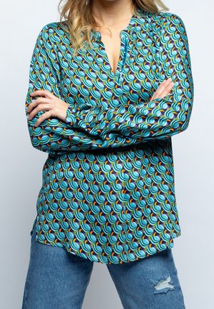 Kvinde iført en teal mønstret bluse med lange ærmer med cirkulære designs og med armene krydset, kombineret med revnede blå jeans.
