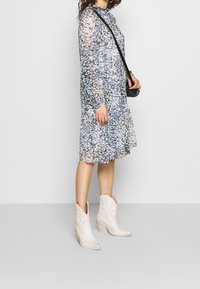 Dlouhá, průhledná šaty s modrou květinovou potiskem, volánkovým výstřihem a dlouhými rukávy. V kombinaci s bílými kovbojskými botami a malou černou kabelkou.