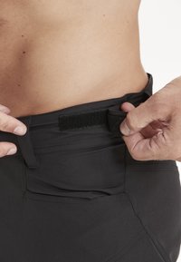 Mustat housut leveällä vyötärönauhalta, joissa on tarrakiinnitys. Suora pinta ja muotoiltu malli, jossa on näkyvät sivutaskut.